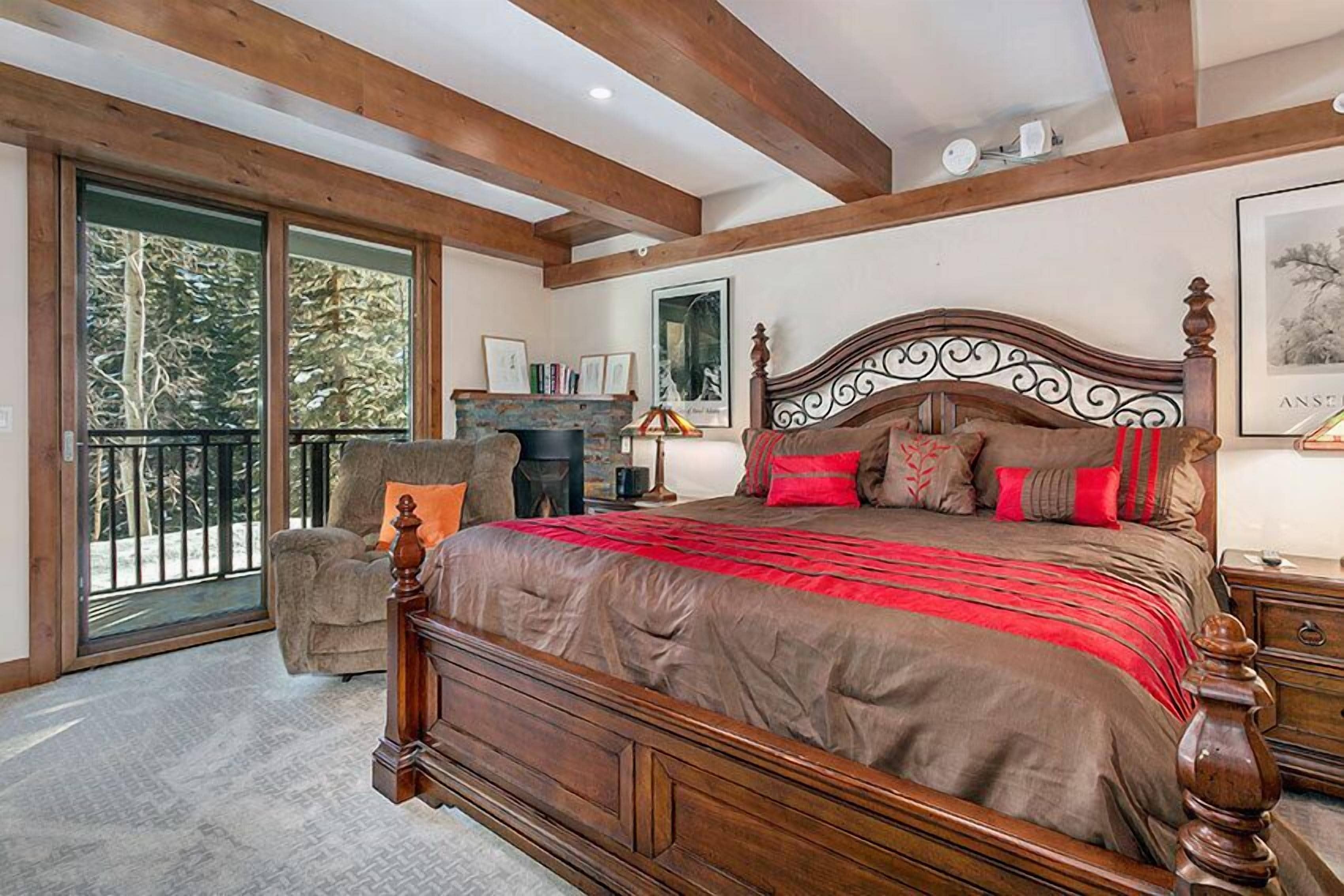 A spacious bedroom with a large bed, wooden beams, a fireplace, and a balcony with a view of trees.