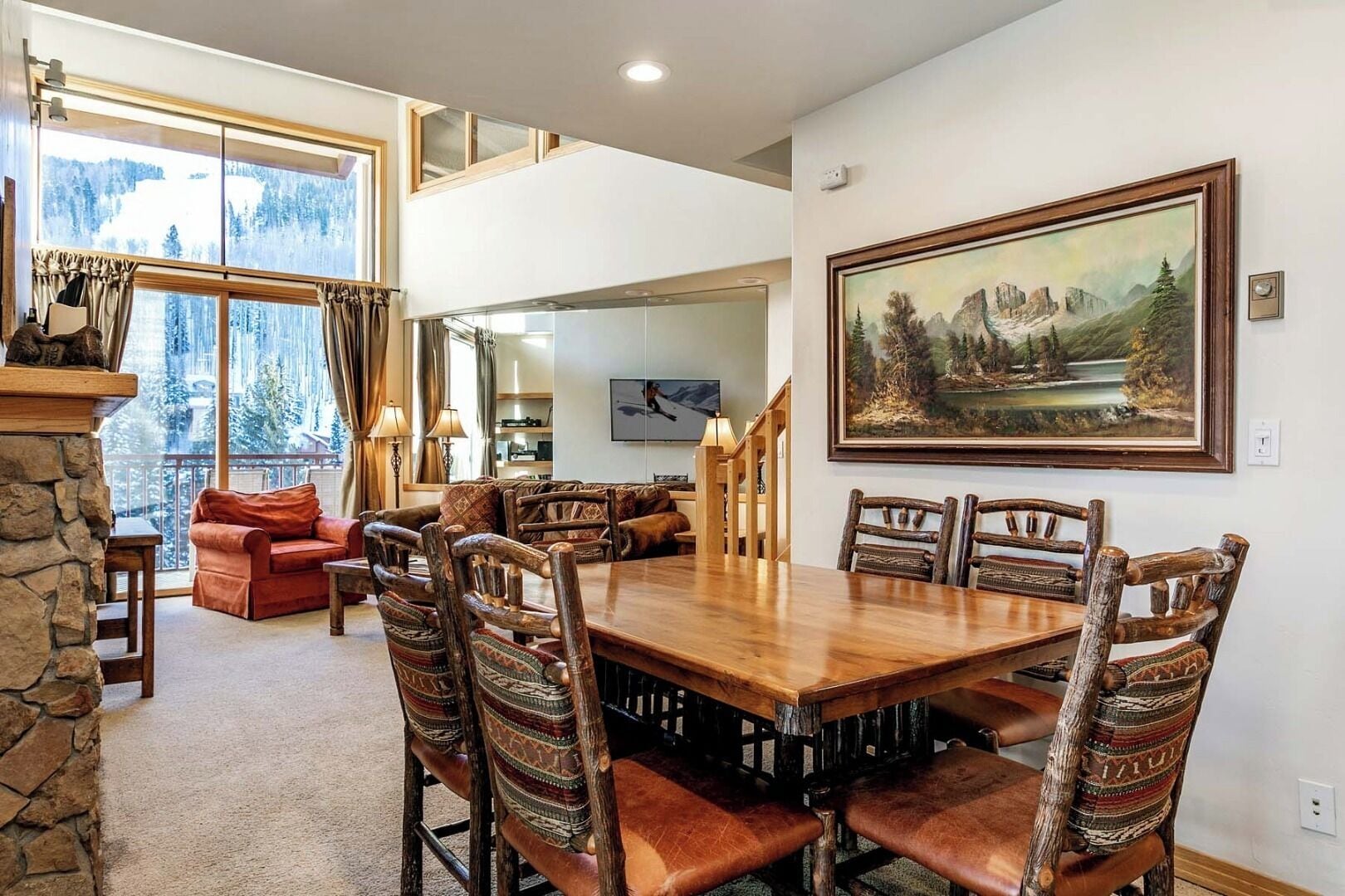 A dining area with a large wooden table and rustic chairs, a fireplace, and a scenic view through large windows.