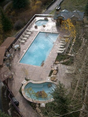 Una piscina rectangular con una piscina circular más pequeña, rodeada de sillas de descanso y mesas, ubicada en un área ajardinada con árboles y un edificio al fondo.