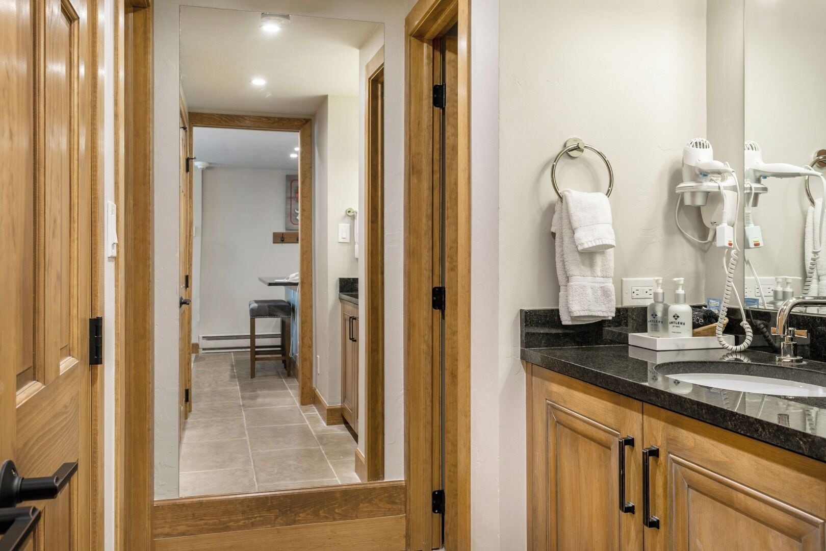 A modern bathroom with a vanity, a sink, and a mirror. There is a towel rack with towels hanging on it, and a hairdryer and other toiletries are visible on the counter.