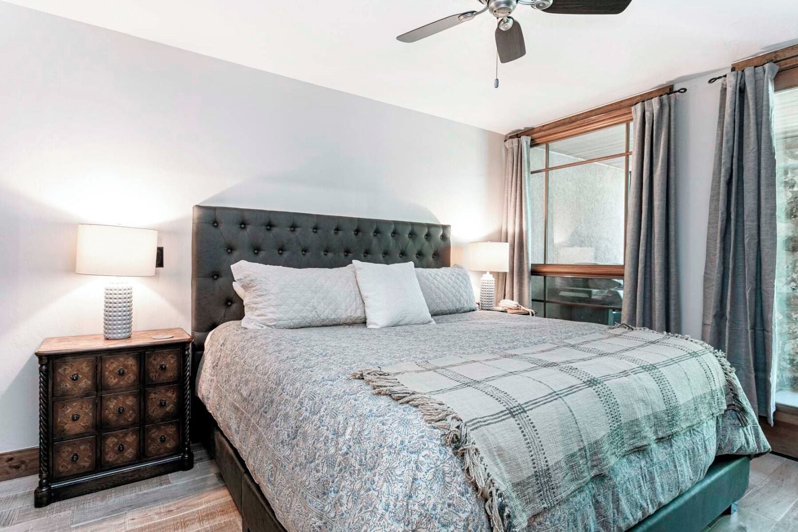 A bedroom with a tufted headboard, a large bed with a patterned blanket, a bedside table with a lamp, and a window with curtains.