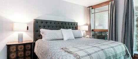 A bedroom with a tufted headboard, a large bed with a patterned blanket, a bedside table with a lamp, and a window with curtains.