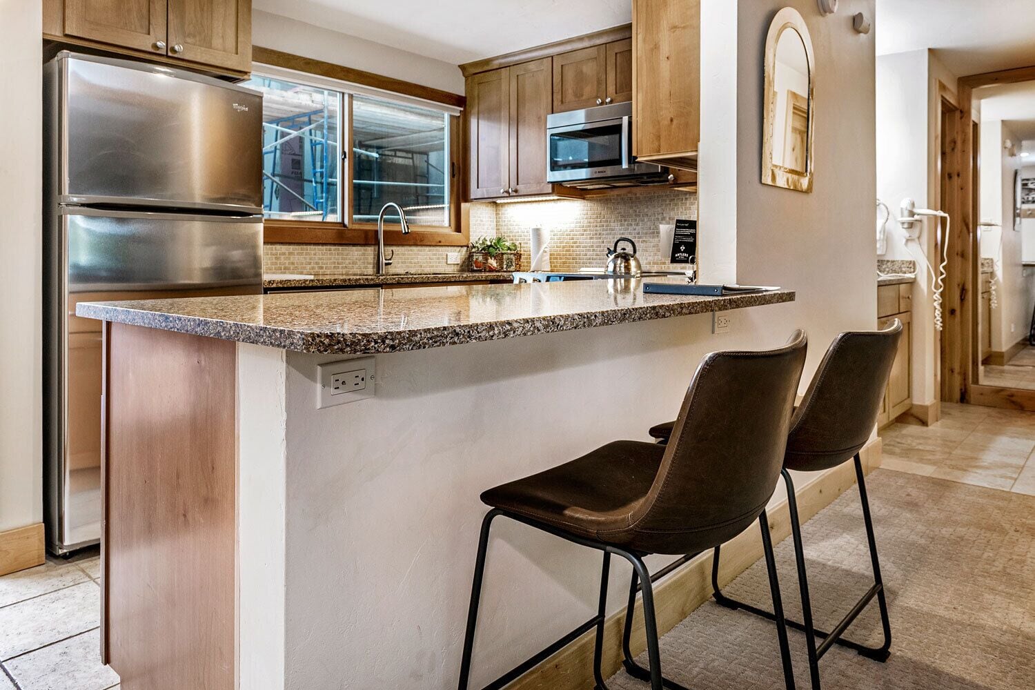A modern kitchen with a stainless steel refrigerator, a microwave, and a sink. There are two bar stools and a window with a view of the outside.