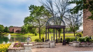A gazebo with a metal roof, surrounded by stone walls and flower beds, near a pond and a brick building.