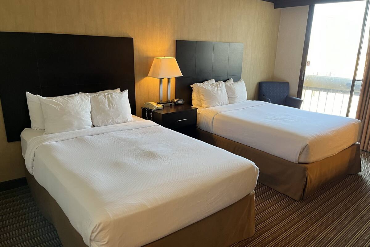 Habitación de hotel con dos camas, una mesita de noche con lámpara y vistas al exterior.