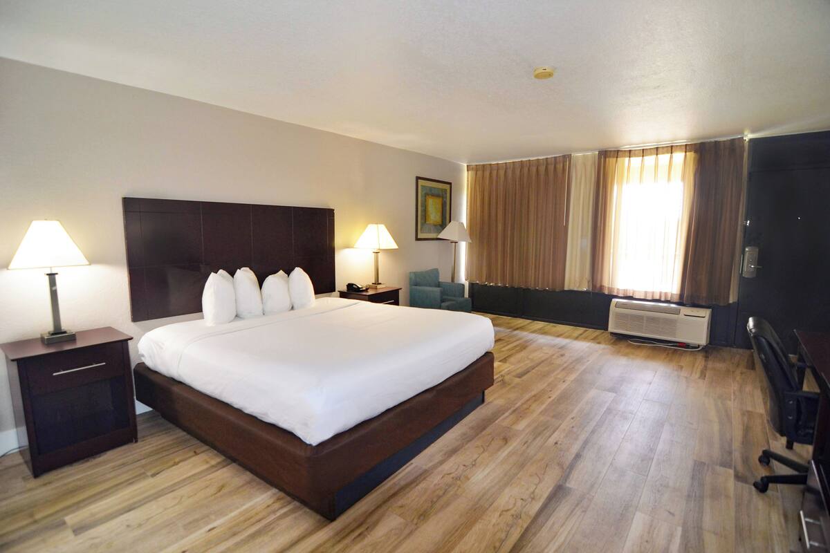 A hotel room with a large bed, two bedside tables with lamps, a desk, a chair, and a window with curtains.