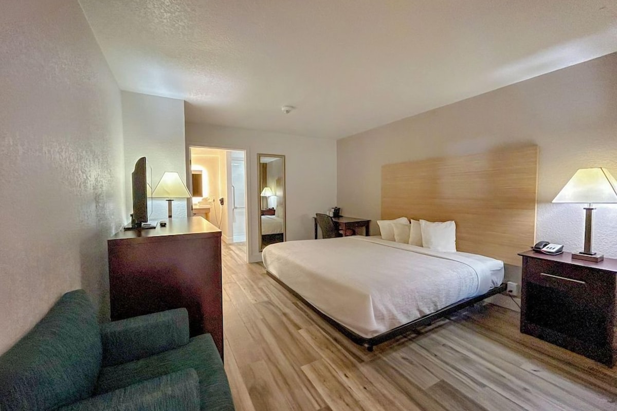 Habitación de hotel con cama, mesita de noche, lámpara y un rincón de estar con una silla.