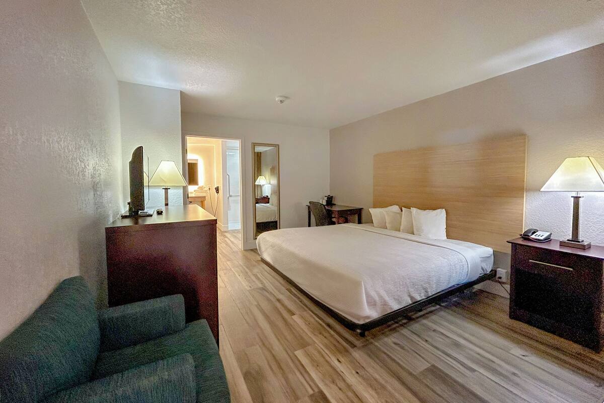 A hotel room with a bed, a desk, a chair, a mirror, a lamp, and a bathroom visible through an open door.