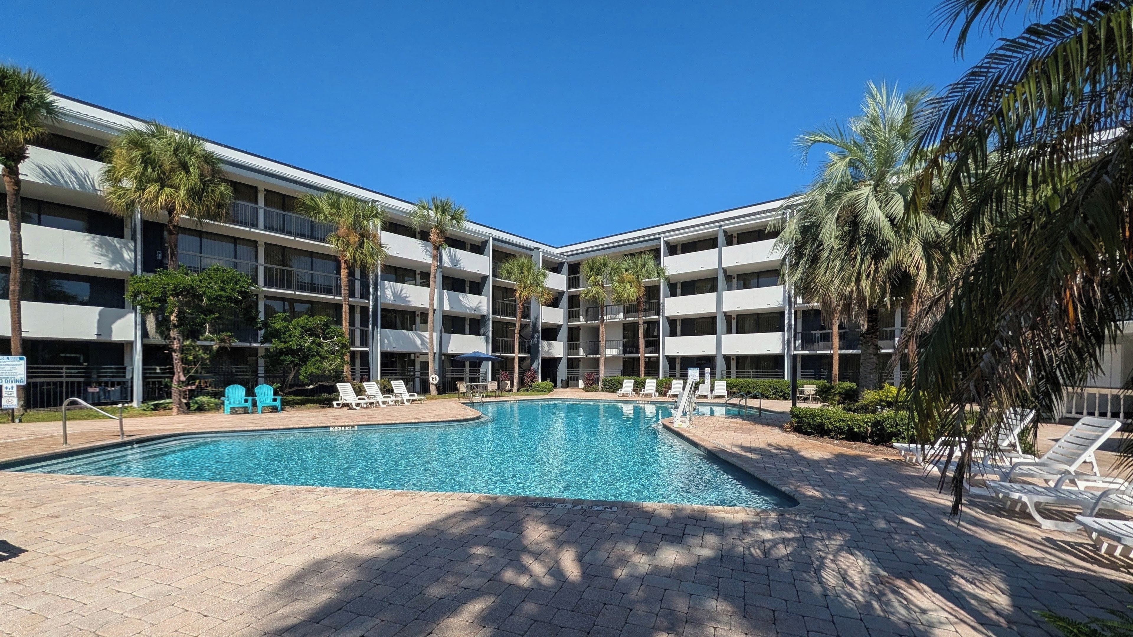 A hotel with a central swimming pool surrounded by lounge chairs and palm trees.