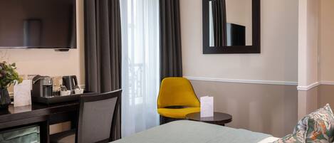 A hotel room with a bed, a desk with a chair, a TV, a mirror, and a window with curtains.