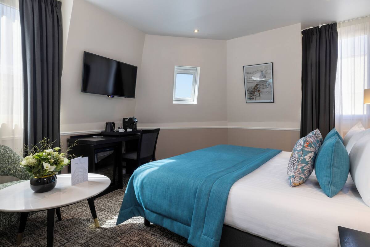 A hotel room with a bed, a desk, a chair, a TV, a window, and a small table with a vase of flowers.