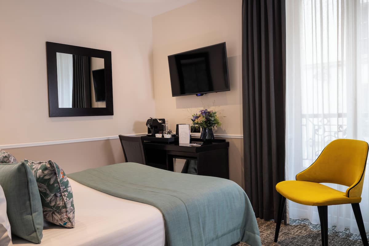 A hotel room with a bed, a desk with a flat-screen TV, a yellow chair, and a window with curtains.