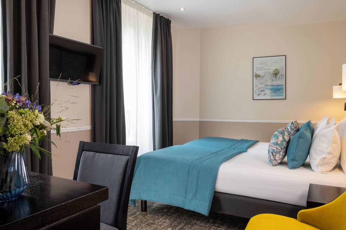 A hotel room with a bed, a desk, a chair, a vase with flowers, and a wall-mounted TV.