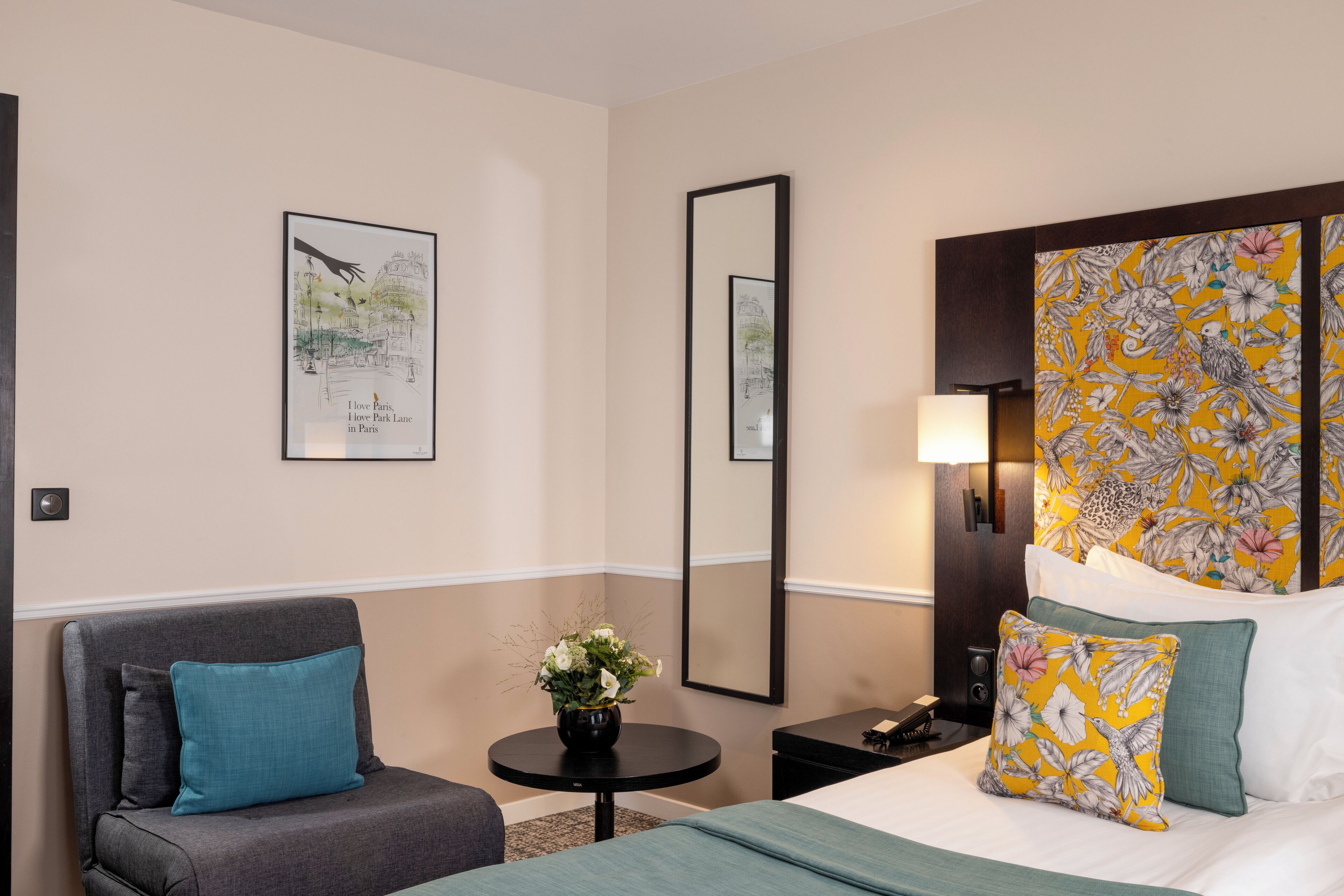 Quarto de hotel com cama, sofá, mesa pequena com um vaso de flores, espelho e quadros nas paredes.