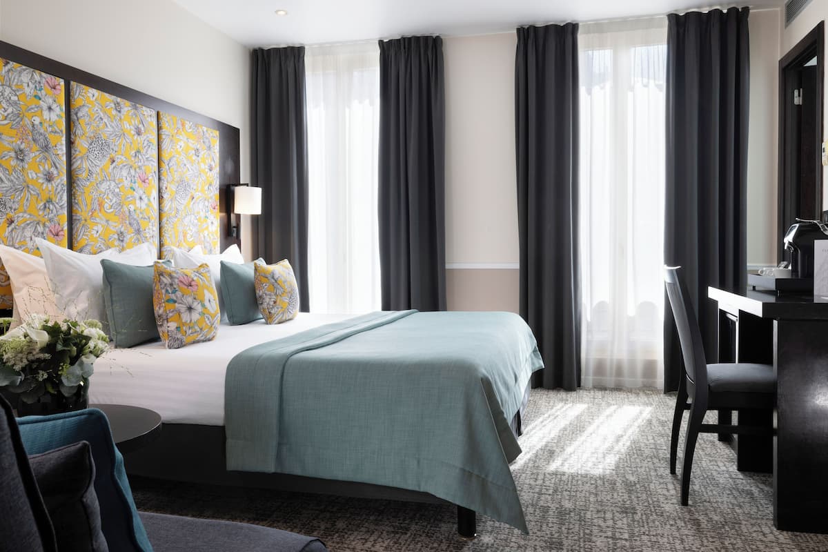 A hotel room with a large bed, a desk, and a chair. There is a window with curtains, a wall-mounted lamp, and a floral artwork above the bed.