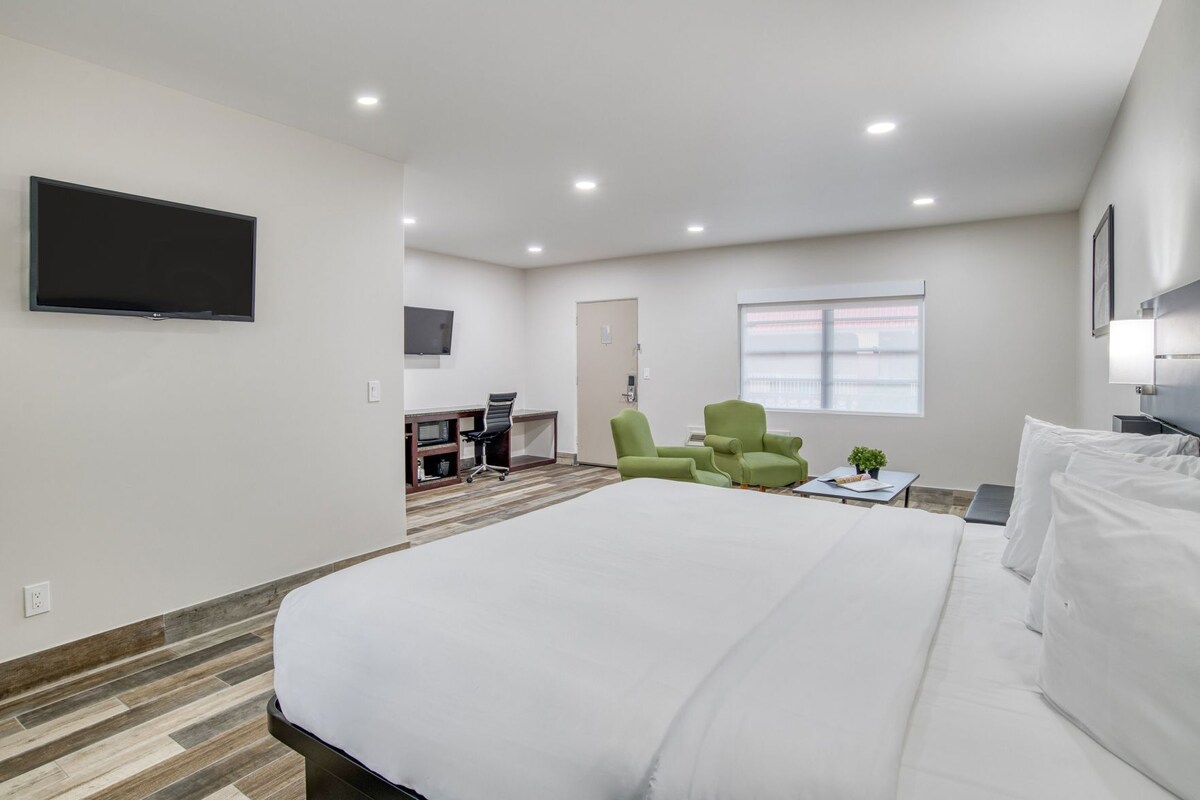 A hotel room with a large bed, a desk with a chair, a green armchair, a TV mounted on the wall, and a window with blinds.