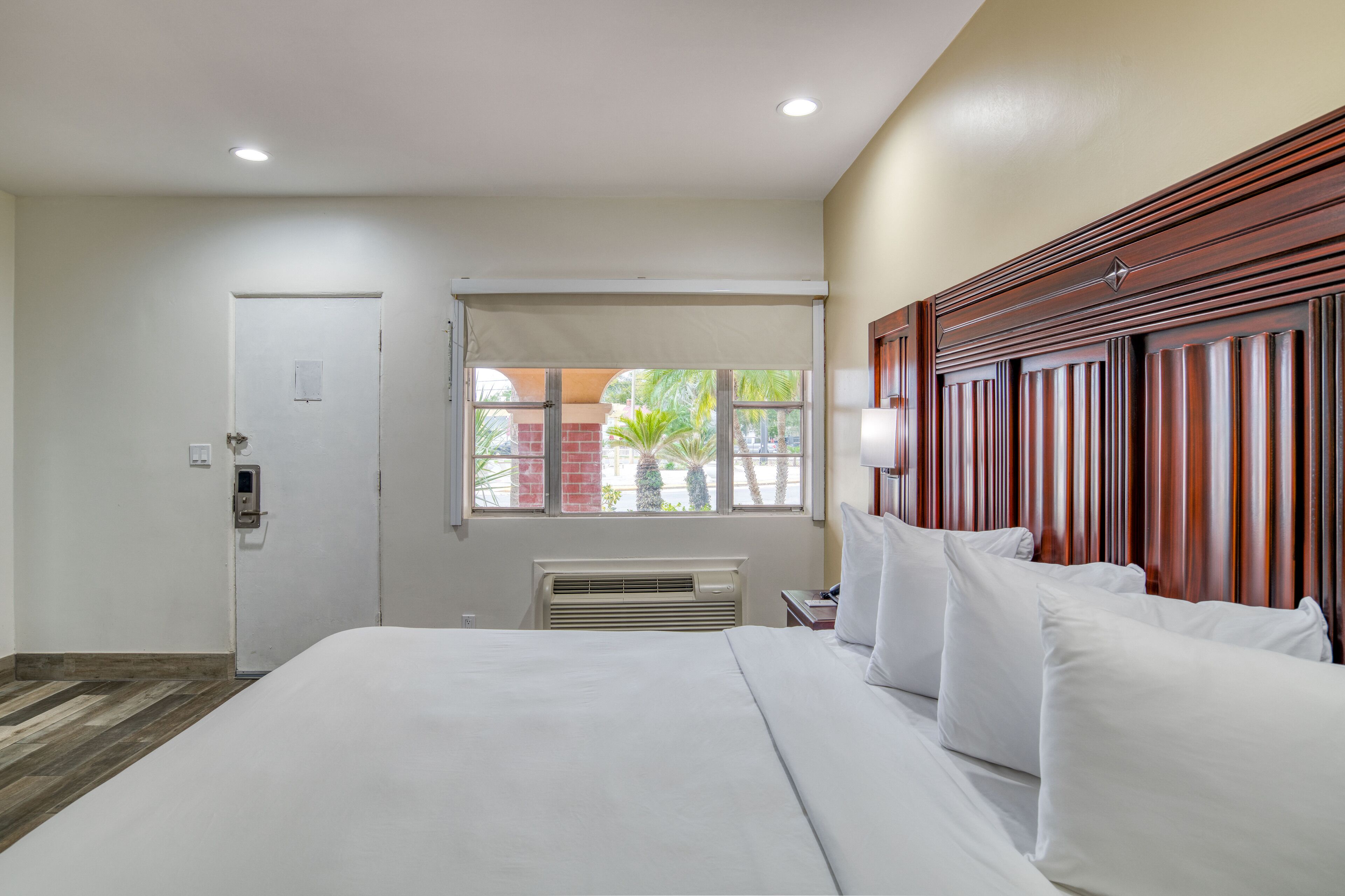 Habitación de hotel con una cama grande, una ventana que ofrece vistas a palmeras, una puerta y un cabecero de madera.