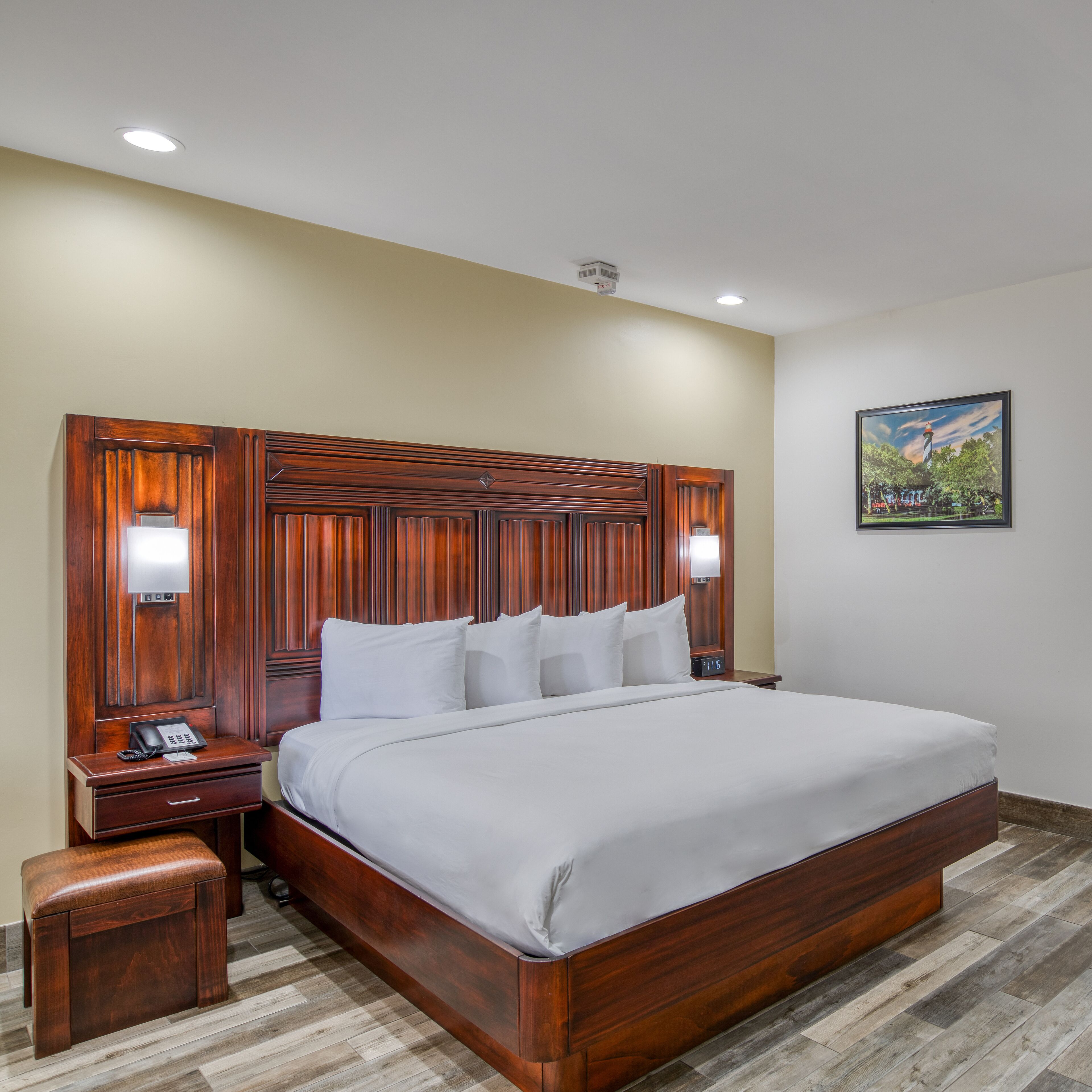 Habitación de hotel con cabecera de madera, cama con sábanas blancas, mesita de noche con teléfono y un pequeño taburete de madera.