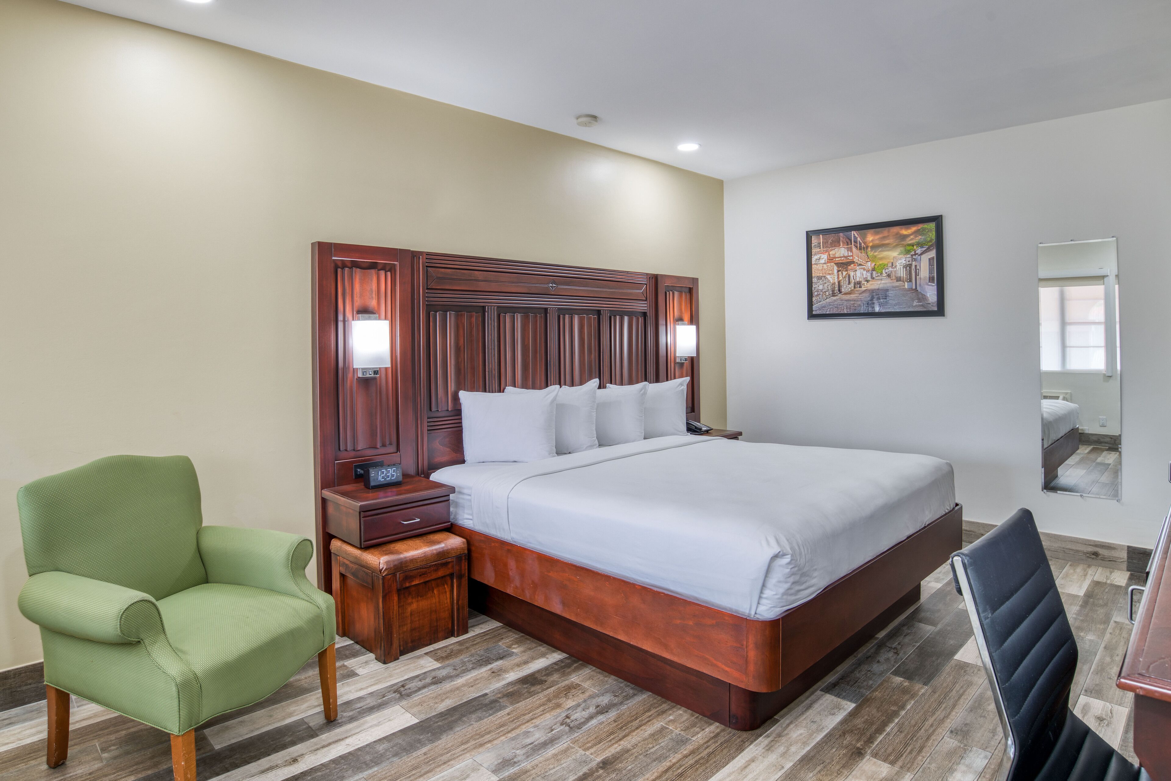 A hotel room with a large bed, a green armchair, a wooden nightstand, and a painting on the wall.