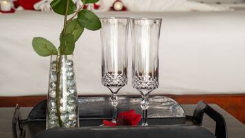 Un plateau élégamment disposé, sur lequel on trouve un vase contenant une seule rose rouge, deux verres en cristal et une rose rouge posée sur un lit agrémenté d’une décoration en forme de cœur, composée de serviettes et d’oreillers blancs.