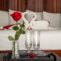 Una bandeja ordenada con un jarrón de una sola rosa roja, dos copas de cristal y una rosa roja sobre una cama con una disposición en forma de corazón de toallas y almohadas blancas.