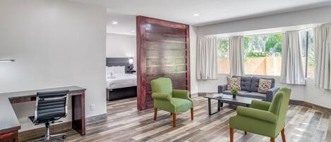 Una habitación de hotel moderna con piso de madera, un escritorio con una silla, dos sillones verdes, un sofá gris y un ventanal con cortinas.