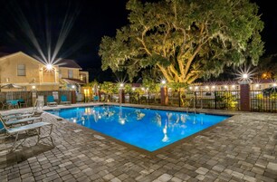 Un área de piscina iluminada por la noche con sillones de descanso, una cerca y un árbol grande.