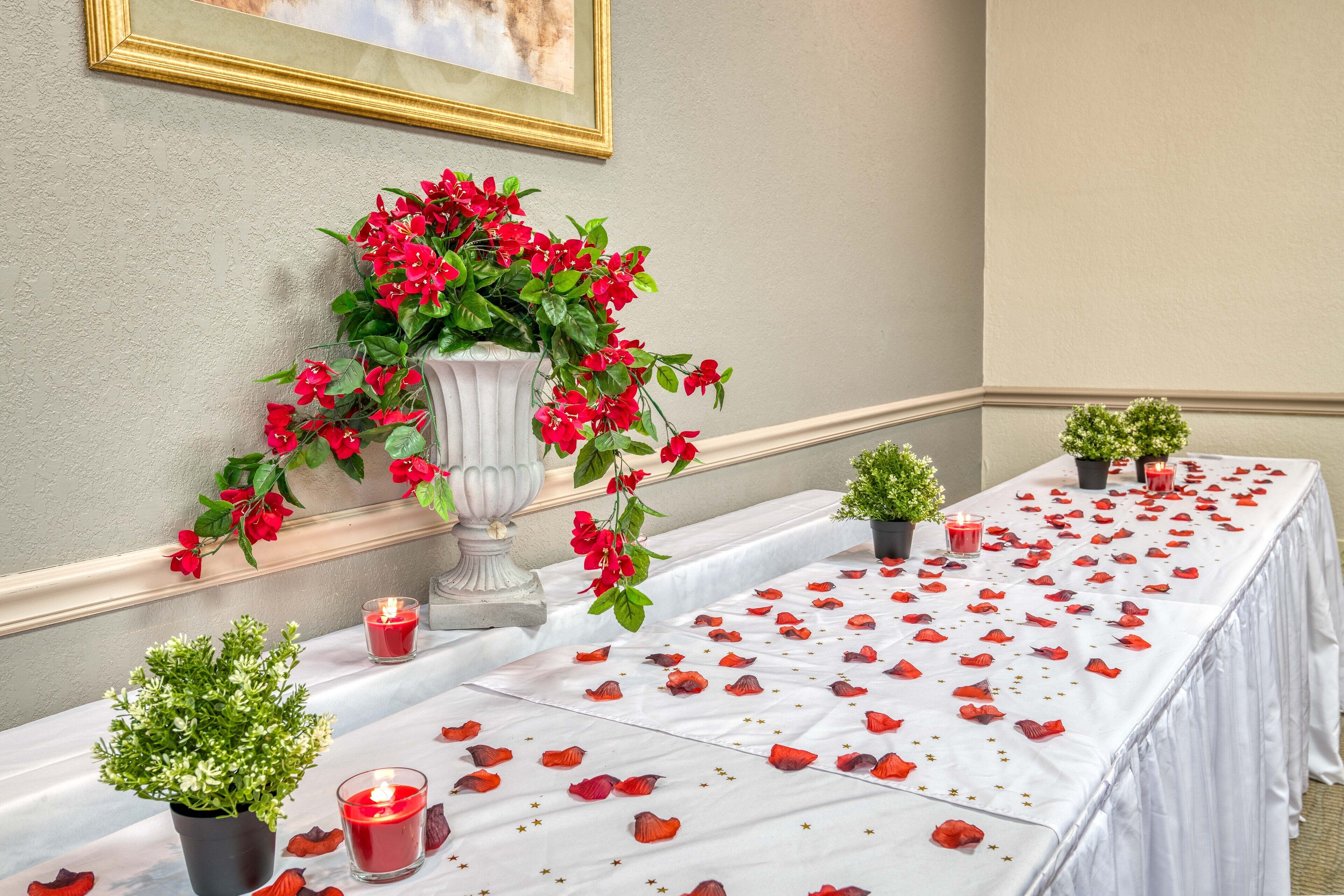 A table set with red rose petals, candles, and small potted plants, suggesting a romantic or special event setting.