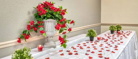A table set with red rose petals, candles, and small potted plants, suggesting a romantic or special event setting.