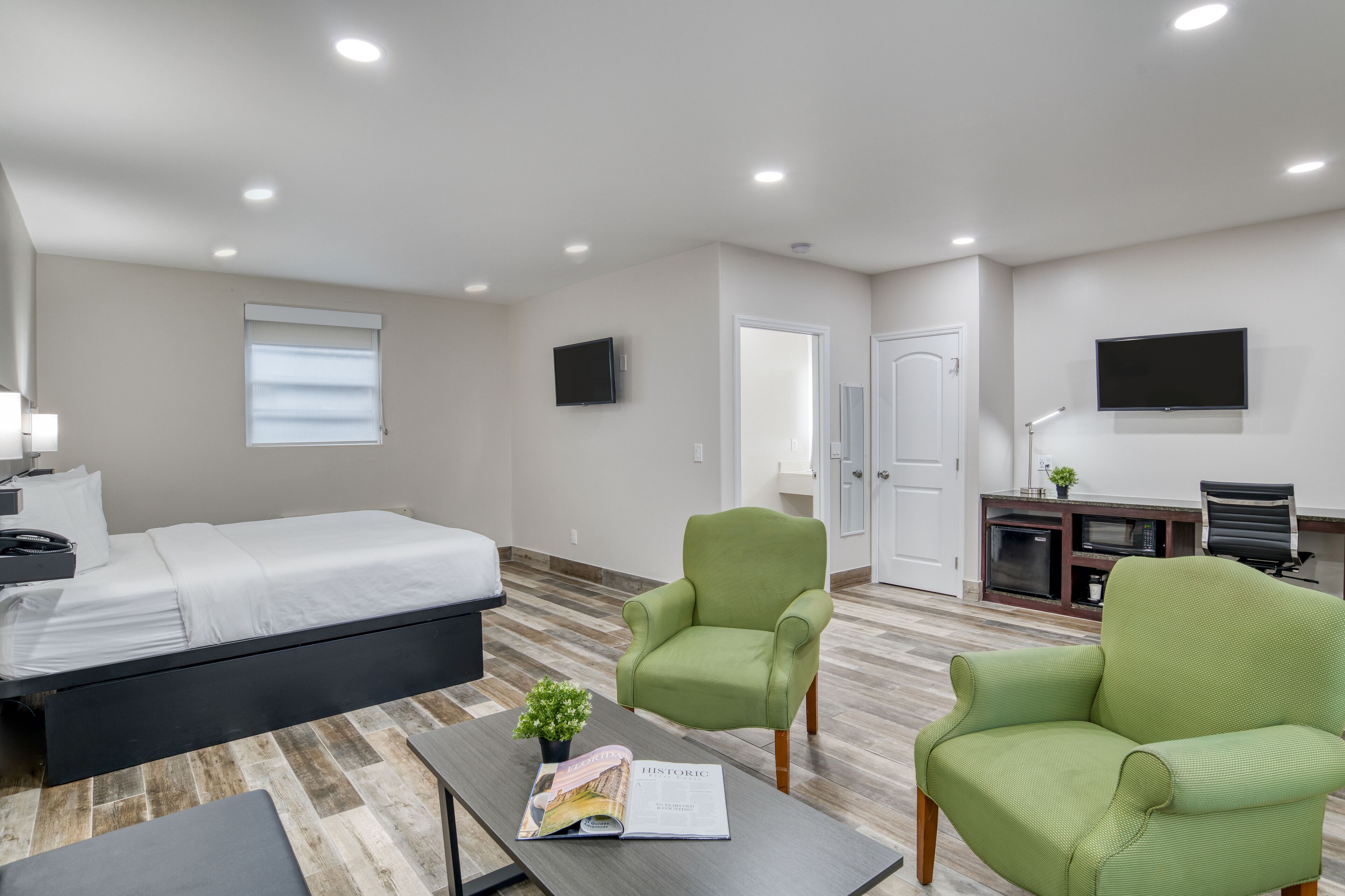 Quarto de hotel moderno com uma cama, duas poltronas verdes, uma escrivaninha com uma televisão e uma pequena planta.