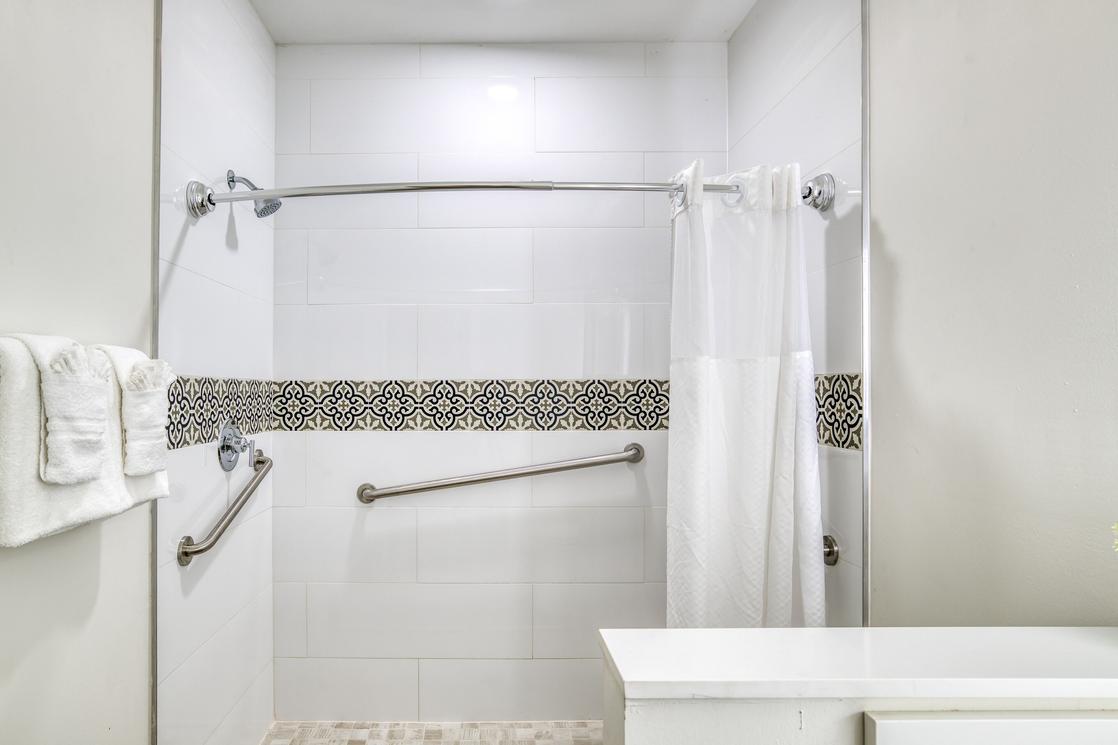A bathroom with white tiles, a shower with a glass door, a towel rack, and a handrail.