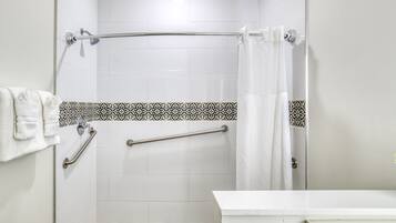 A bathroom with white tiles, a shower with a glass door, a towel rack, and a handrail.