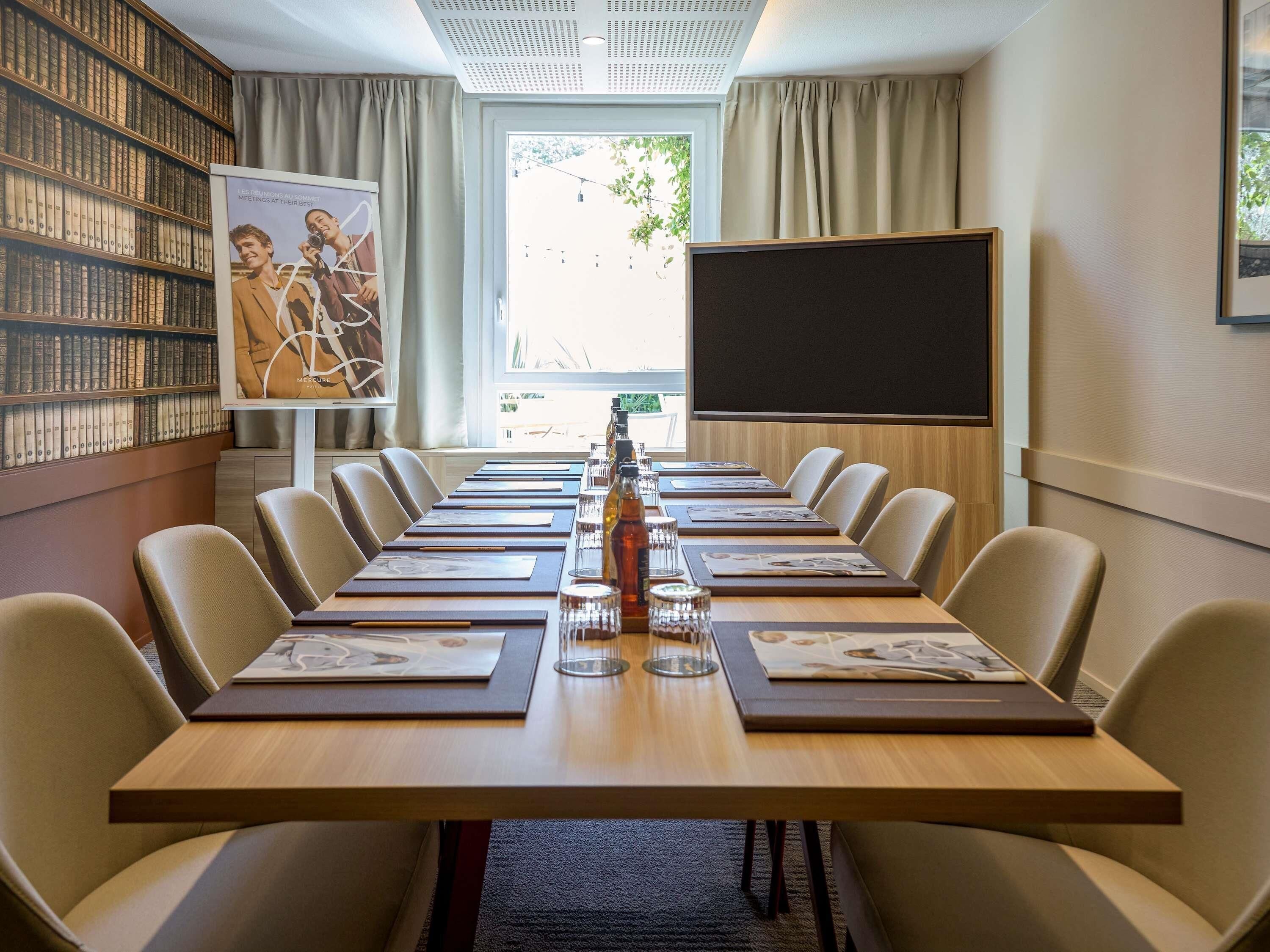 A conference room with a long table, chairs, a projector screen, a bookshelf, and a window with curtains.