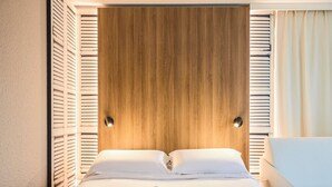 Habitación de hotel con cabecera de madera, dos almohadas blancas, cubrecama blanco, una silla blanca y una bata blanca sobre la cama.