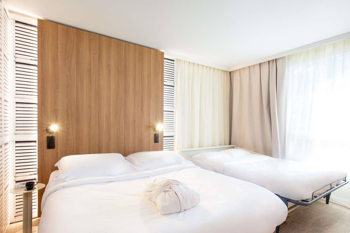 A hotel room with two beds, a wooden accent wall, a bedside table with a lamp, and a window with curtains.