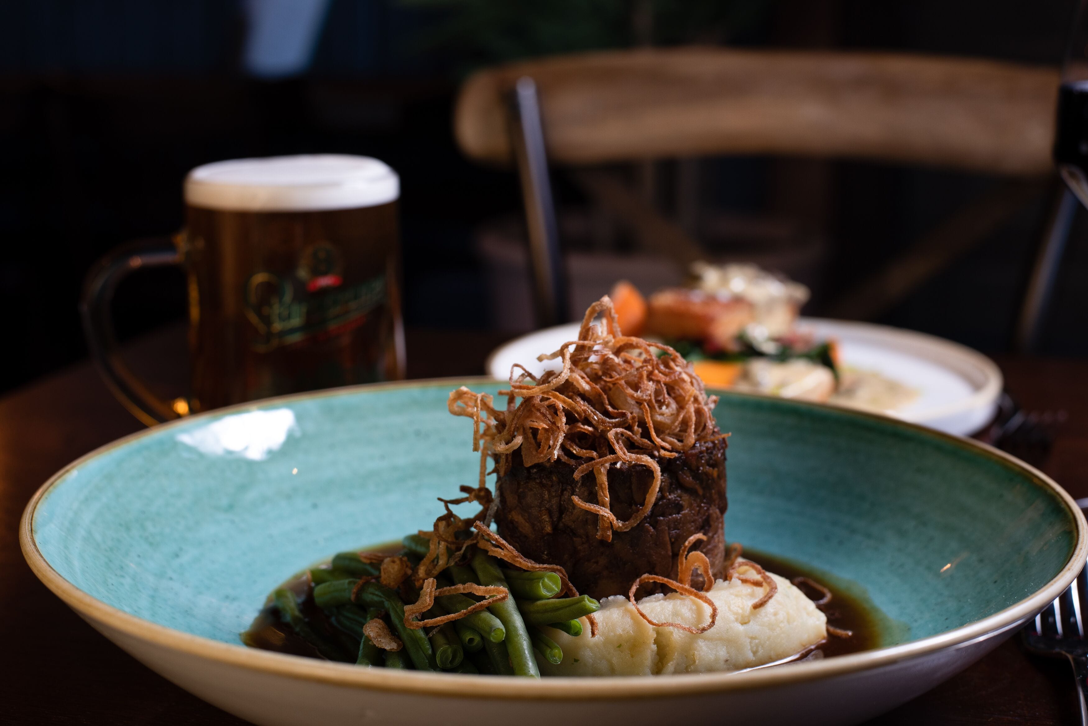 A plate of food with a mound of mashed potatoes, green beans, and a beef steak topped with fried onions, accompanied by a mug of beer.
