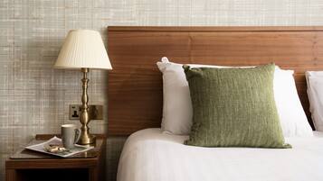 A neatly made bed with white linens and a green pillow, a bedside table with a lamp, a mug, and a small item on the table.
