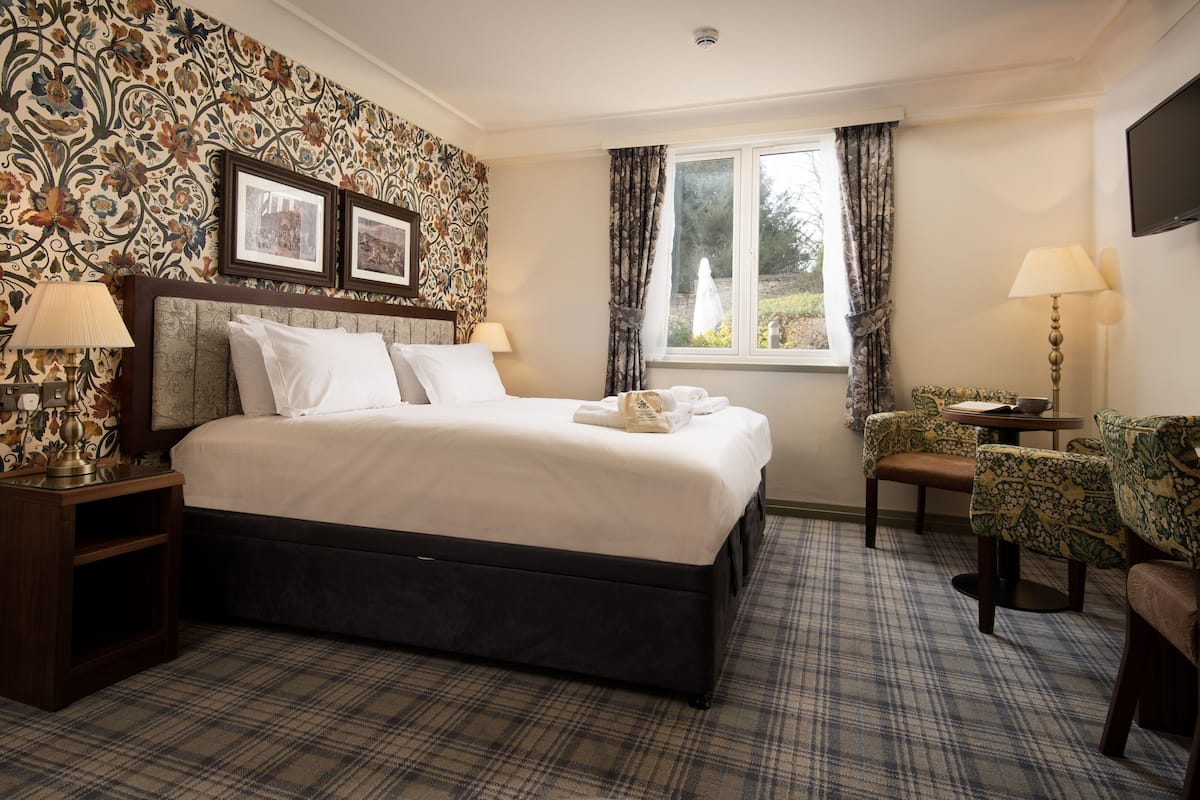 A hotel room with a large bed, two bedside tables with lamps, a window with curtains, a small table with chairs, and a wall with patterned wallpaper.