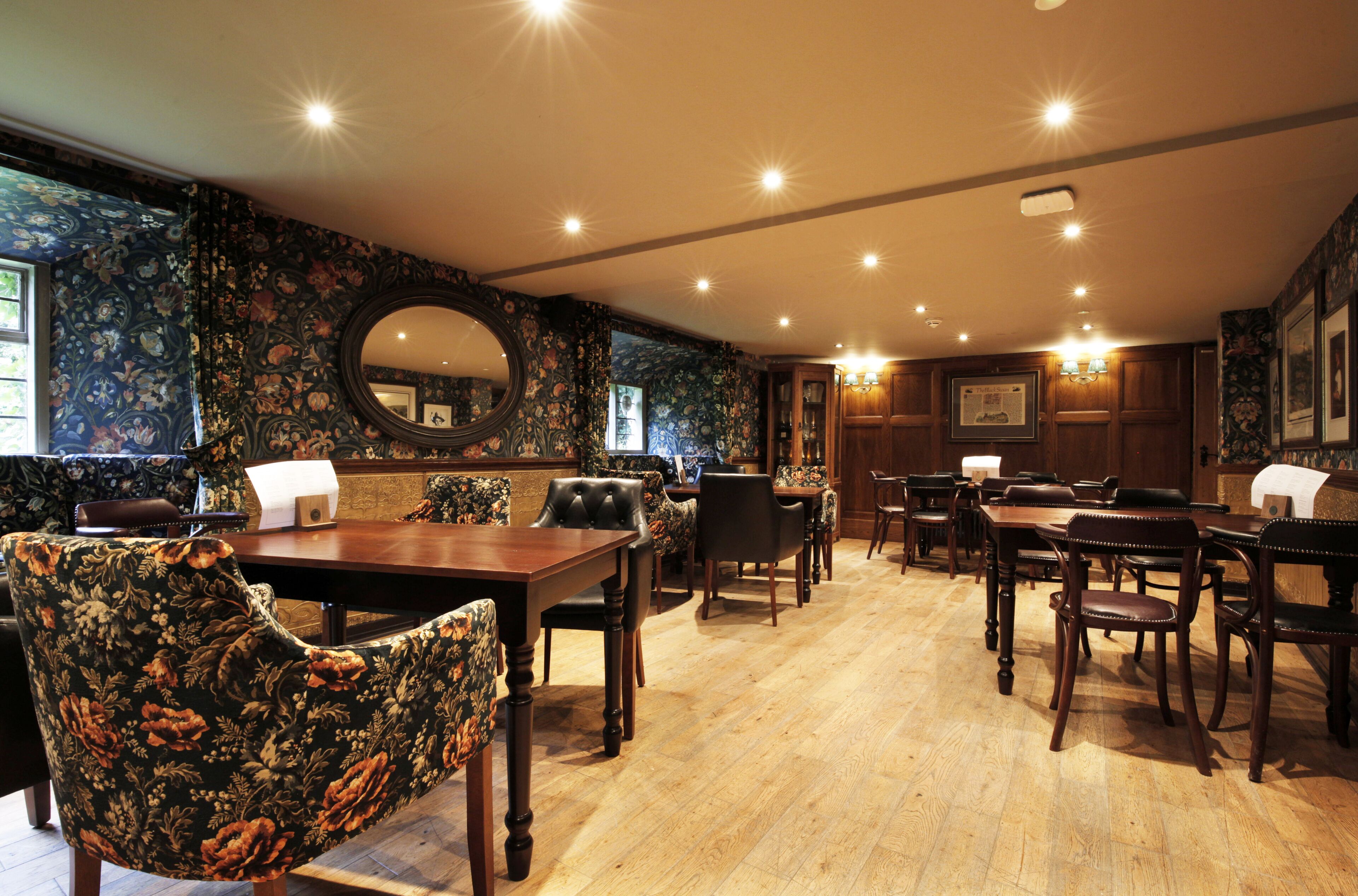 A dining area with wooden tables and chairs, floral wallpaper, and a large mirror.