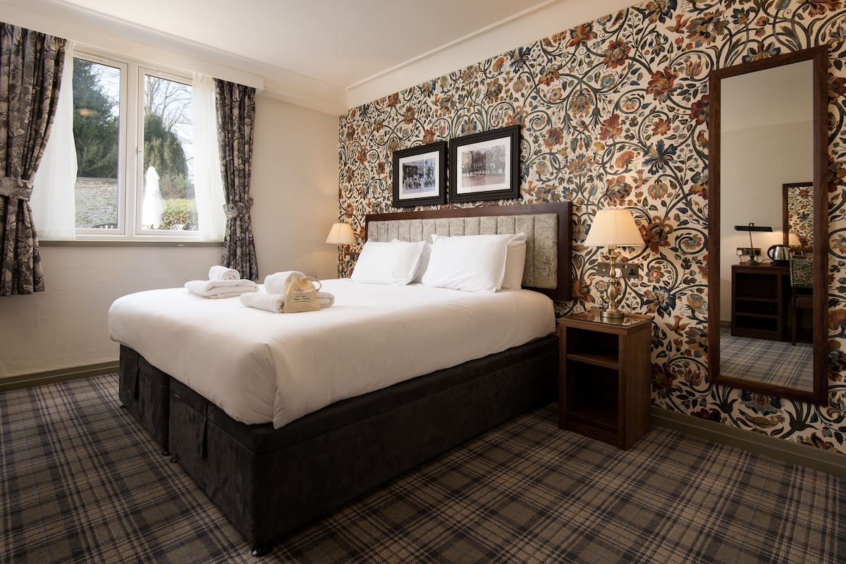 A neatly made bed with white linens, a dark headboard, and a plaid carpet. There are two bedside lamps and a mirror on the wall.