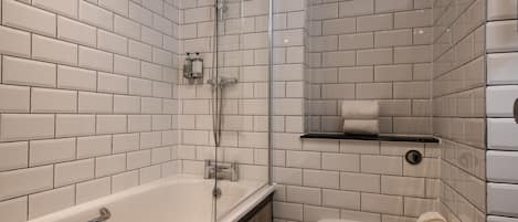 A modern bathroom with white subway tiles, a glass-enclosed shower, a white bathtub, and a toilet with a paper towel holder.