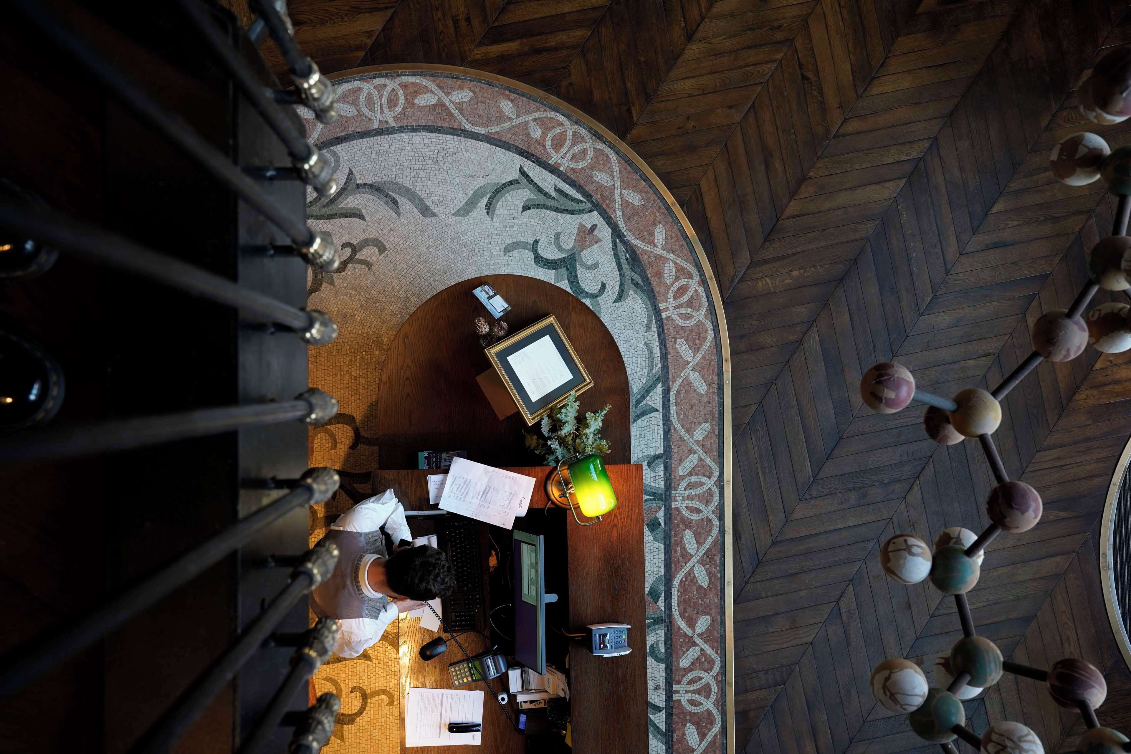 A room with a desk, computer, and various papers, with a patterned carpet and wooden ceiling.
