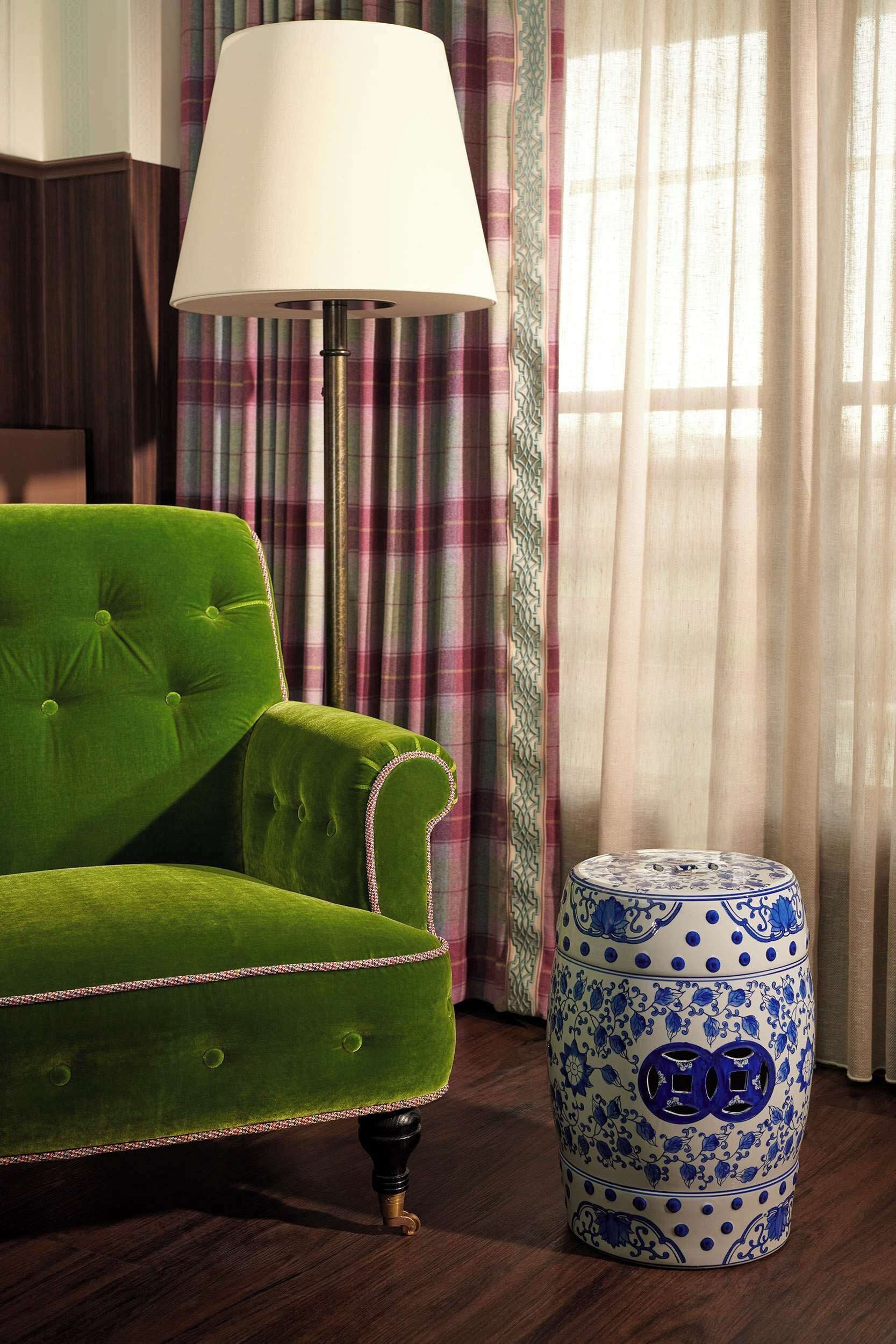 A green armchair, a white floor lamp, and a blue and white patterned ottoman in a room with striped curtains and wooden flooring.