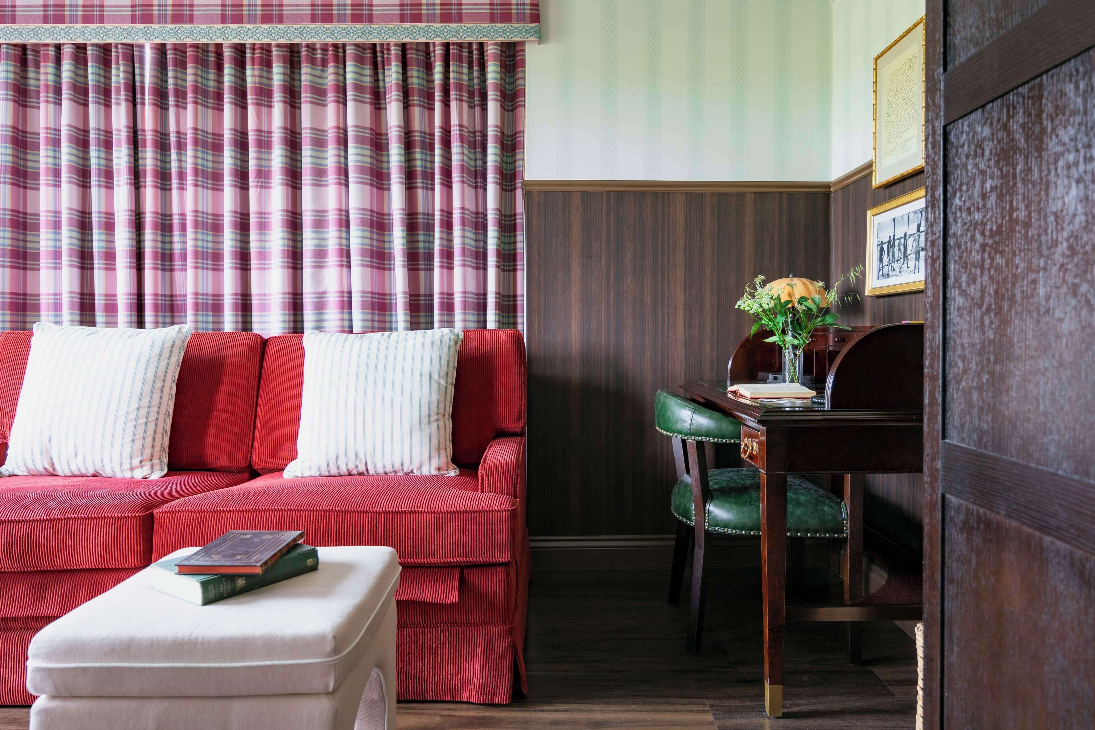 A room with a red sofa, a green chair, a small table with a vase, and a white ottoman.