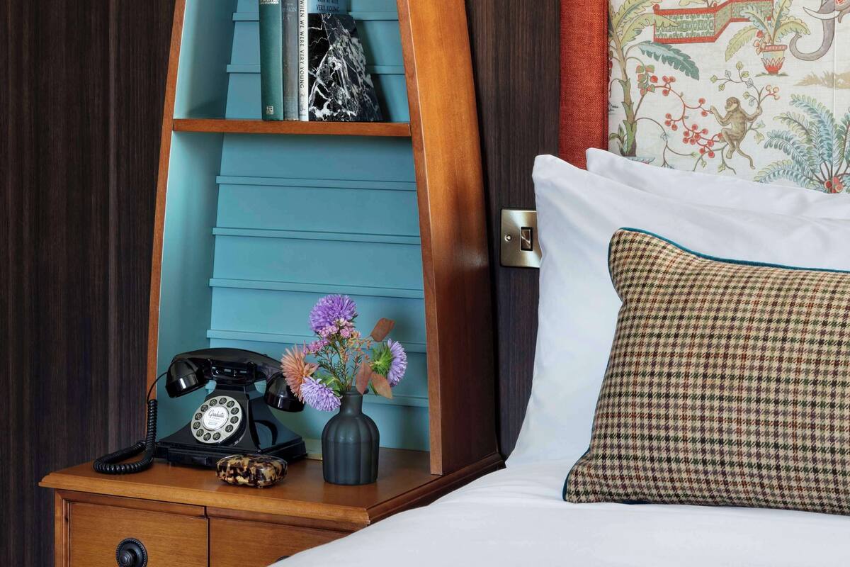 A room with a bed, a nightstand with a telephone and a vase of flowers, a bookshelf with books and a lamp, and a patterned headboard.