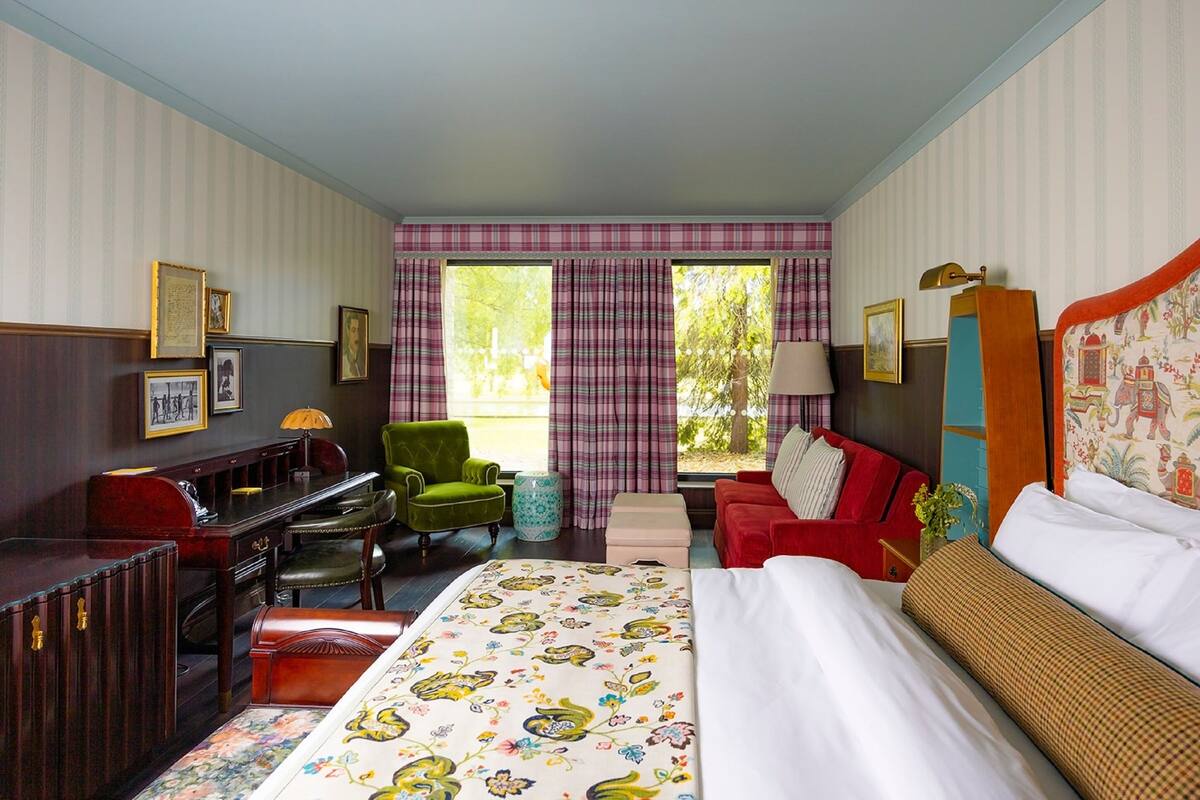 Ett hotellrum med en säng, ett skrivbord, en grön fåtölj, en röd soffa och ett fönster med gardiner.