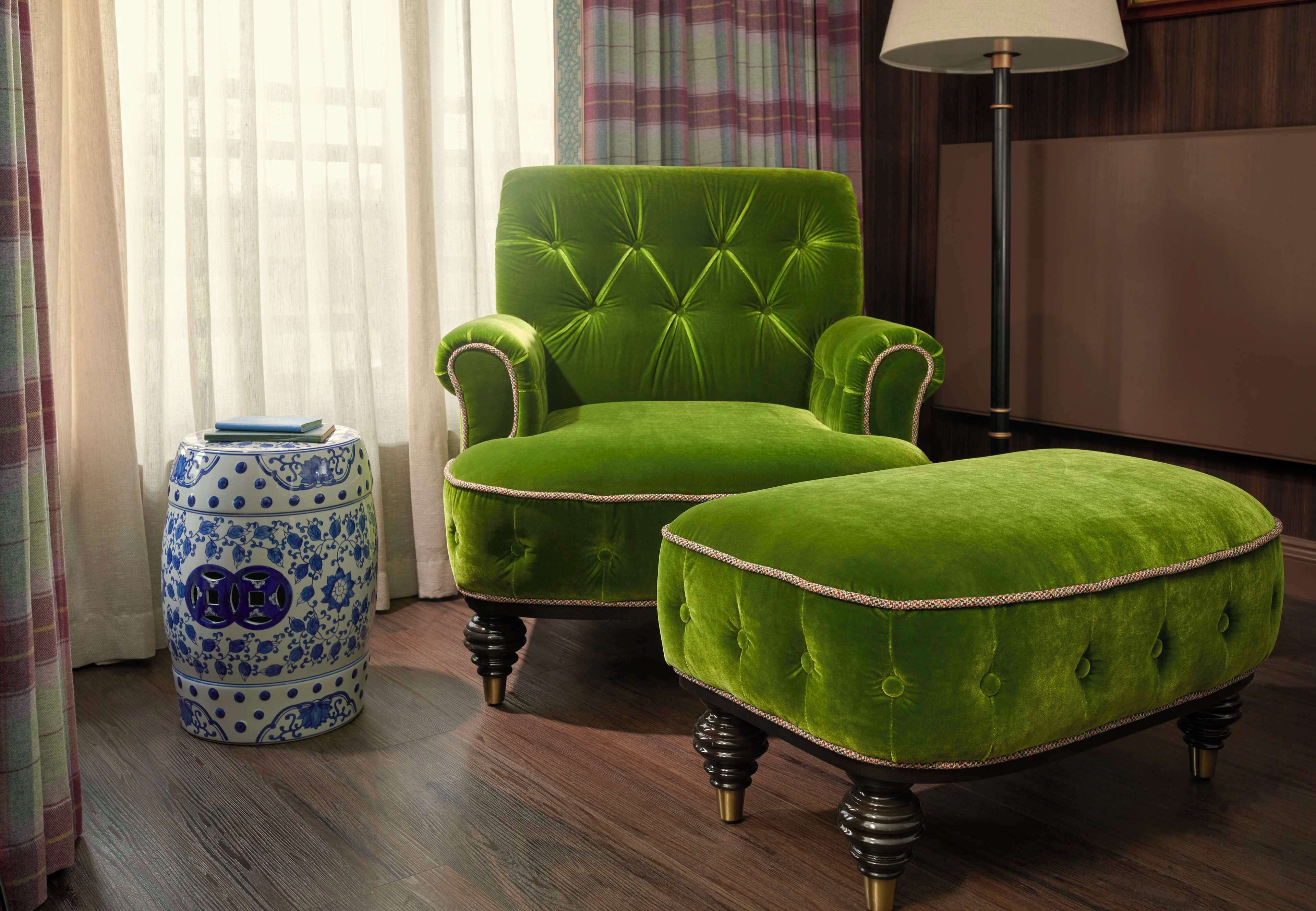 A green upholstered armchair and ottoman with a tufted backrest and armrests, positioned next to a blue and white ceramic vase with floral patterns.