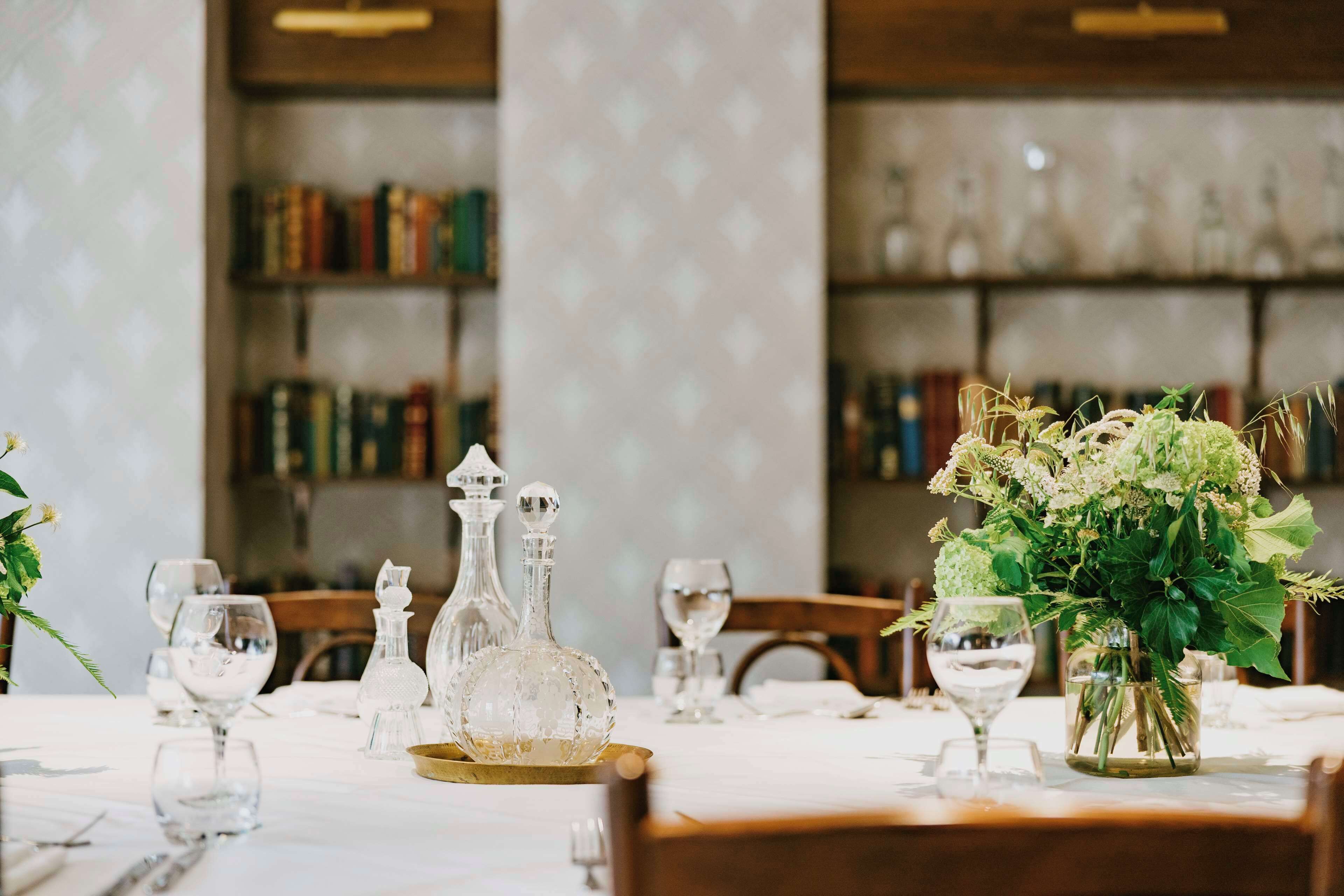 A dining table set with crystal glassware, a vase with flowers, and a bookshelf in the background.