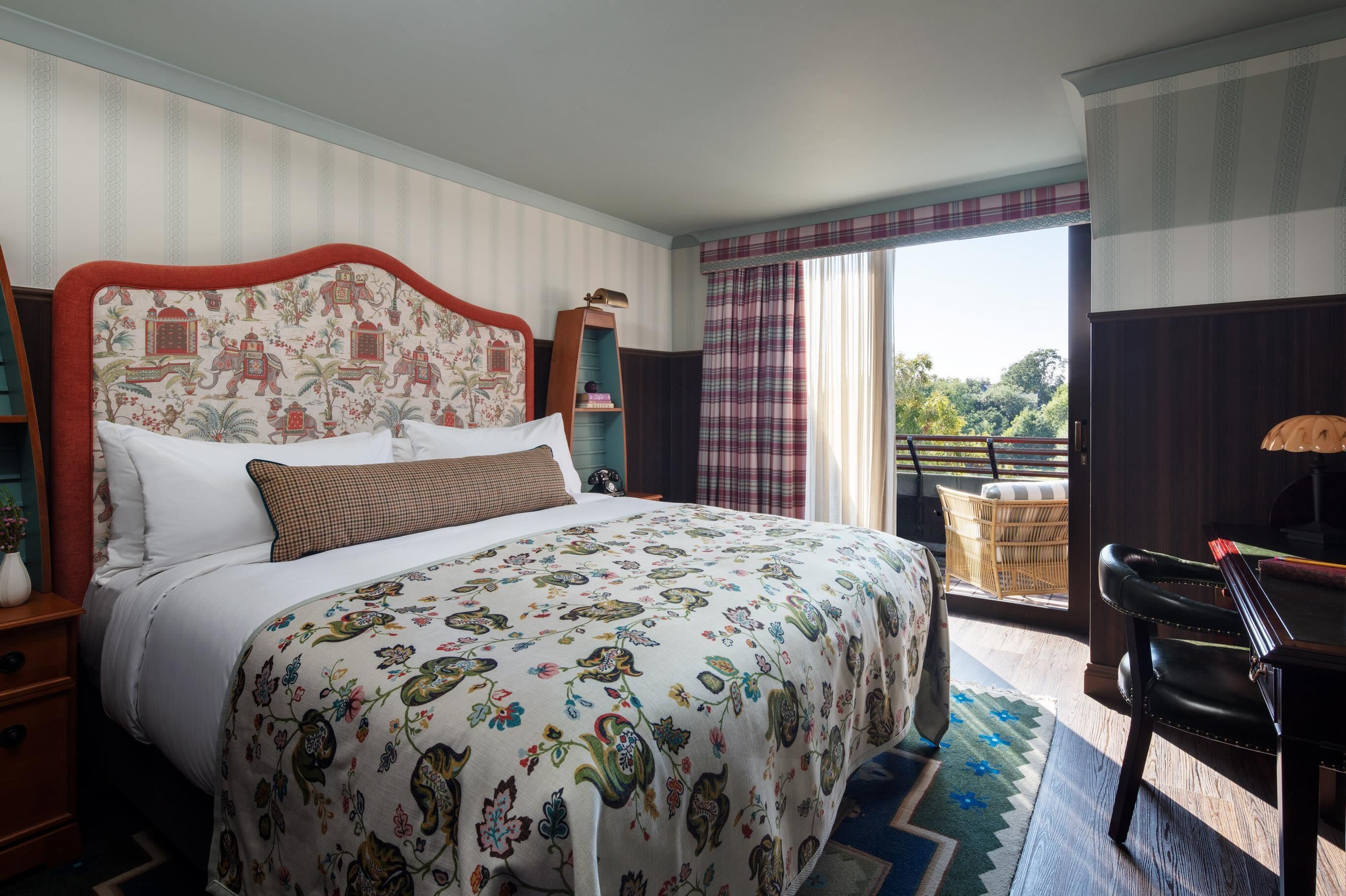 A hotel room with a large bed, a wooden nightstand, a desk with a chair, a wicker chair, and a view of the outdoors through a window with curtains.