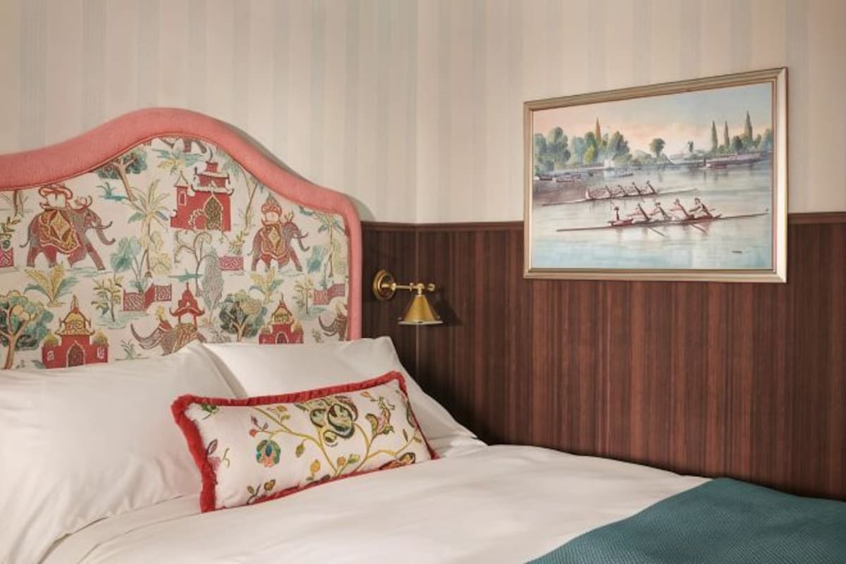 A bed with a patterned headboard, a painting of rowers on a river, and a wall-mounted lamp.